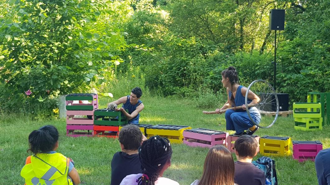 Zwei Schauspielerinnen spielen auf der Gartenbühne vor Kindern ihr Stück. Eine Schauspielerin hält eine Igel-Handpuppe in der Hand, die andere einen alten Fahrradreifen.
