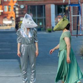 Zwei Schauspieler tragen jeweils eine Maus-Maske und sehen sich an. Eine trägt ein grünes Kleid, eine einen silbernen Anzug.