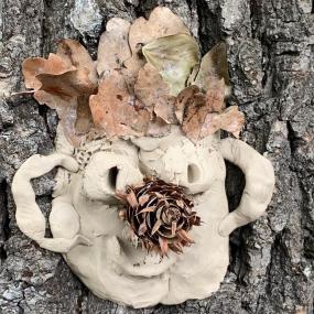 Ein Gesicht aus Ton an einem Baumstamm. Das Gesicht wurde mit Stöckern, Blättern und Blüten verziert. 