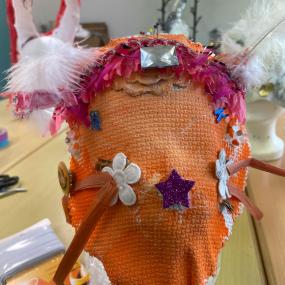 Eine orangene Maske mit Verzierungen darauf und zwei Hörnern aus Federn. 
